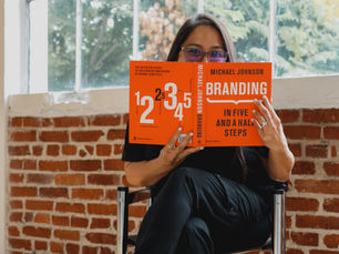 Woman wearing black and white sneakers sitting playfully holding a branding book that covers her face. 