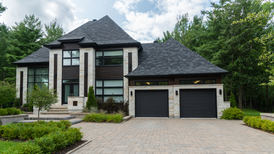 Modern house with dark roof, stone facade, and large windows. Two black garage doors. Surrounded by green trees and shrubs on a cloudy day.