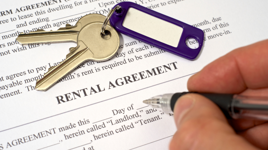 Keys with a purple tag rest on a rental agreement. A hand holding a pen appears ready to sign. The mood is formal and businesslike.