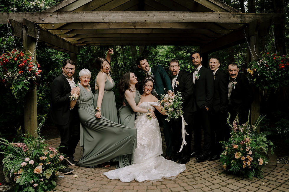 Wedding party poses playfully under a wooden gazebo. Bride in white, others in green and black. Lush greenery and flowers surround them. Dark and moody romantic wedding Vancouver photographer.