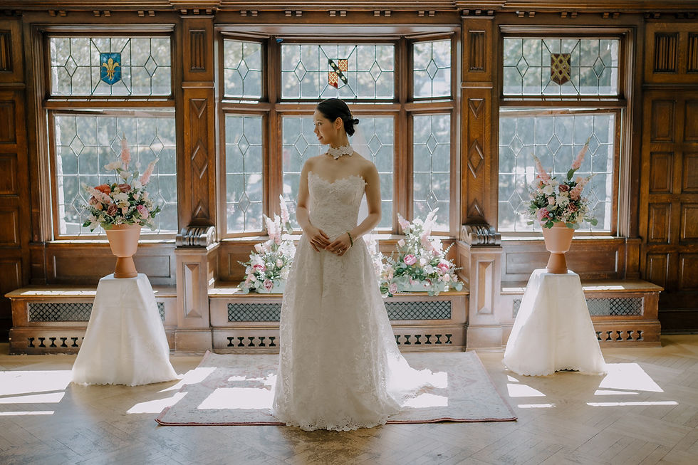 Bride in a lace white gown stands in a wood-paneled room with stained-glass windows, framed by pink-and-white floral arrangements. Aberthau Mansion in Vancouver, Heartfelt, moody and timeless Wedding Photography.