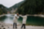 A man kisses a woman's hand by a serene lake, surrounded by dense green forests and mountains. Both wear light coats, creating a romantic mood. Dark, moody and romantic wedding photography.