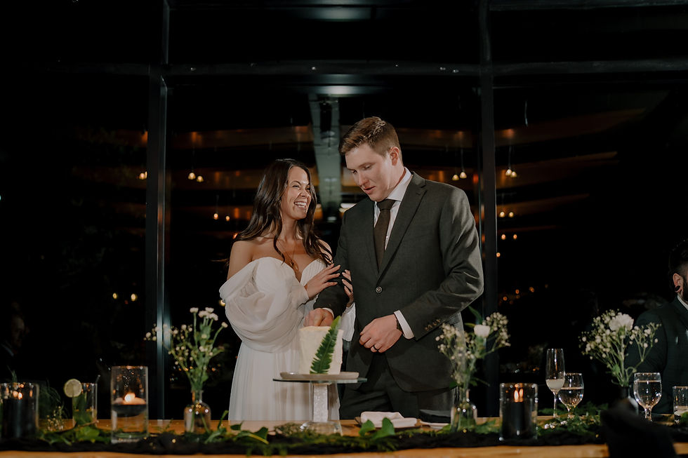 A couple smiles while cutting cake at a table set with candles and flowers in a dimly lit room with sparkling lights in the background.  Vancouver Moody Romantic intimate Wedding Photographer.
