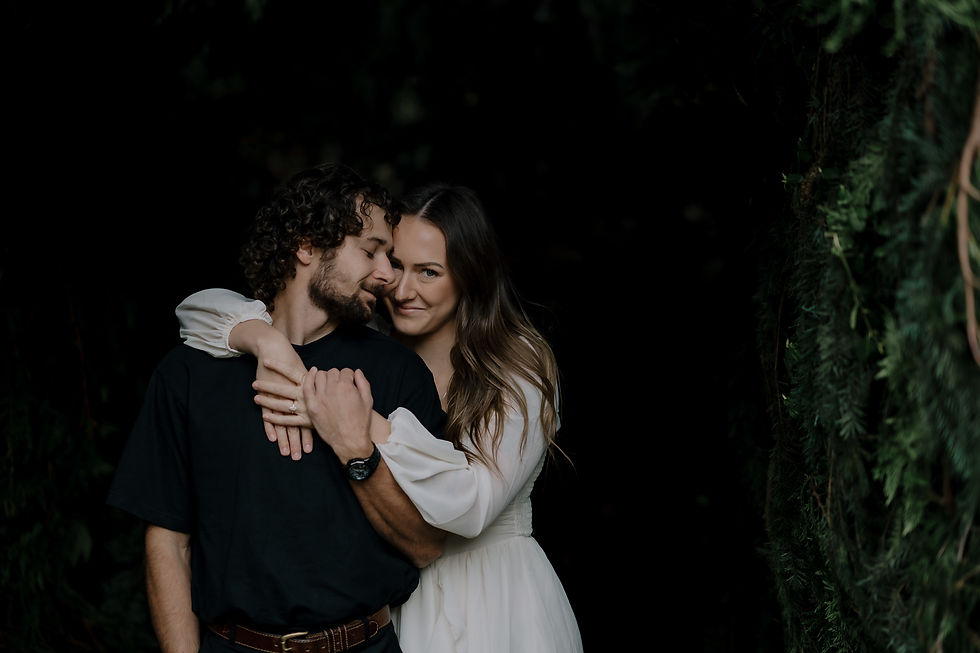 A couple embraces warmly, the woman in a white dress, the man in a dark shirt. They stand amidst lush greenery, exuding a tender mood.
