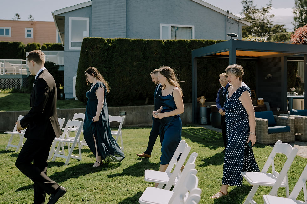 Well-dressed guests walk between white chairs on a sunny lawn during an outdoor ceremony beside a house and patio pergola. Moody and intimate Vancouver wedding photographer with a dark and heartfelt signature look. Small backyard wedding documented.