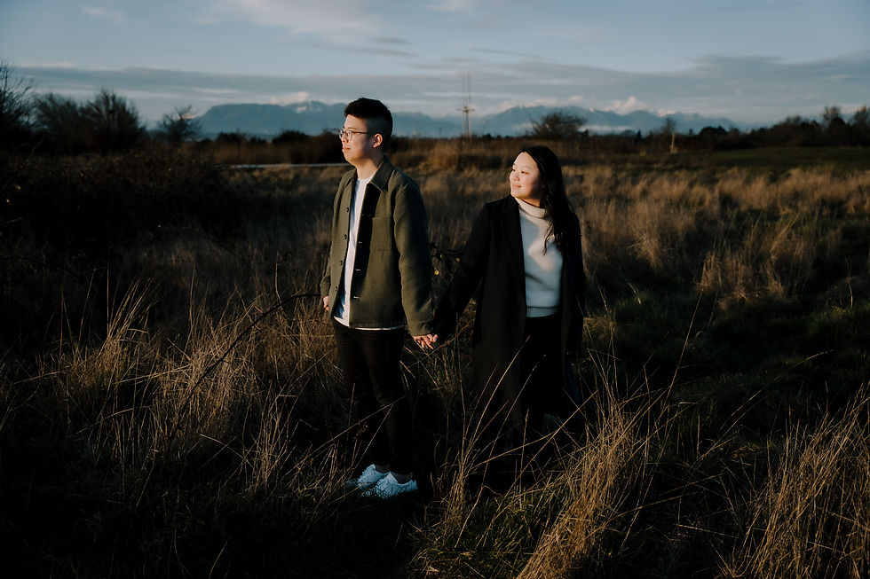 A couple holds hands in a grassy field, looking in different directions. The sky is partly cloudy, and the atmosphere is serene. candid photography session