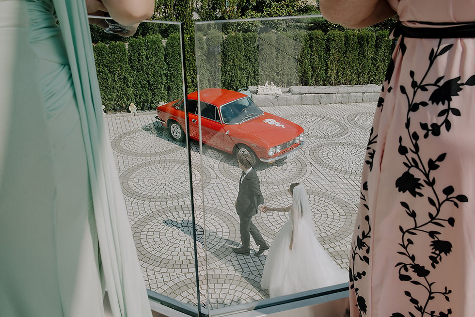 Bride and groom hold hands in a tiled courtyard, framed by hanging dresses, beside a red vintage car and green hedges. This Alfa Romero car added style to this epic wedding. Intimate, moody, cinematic Wedding photography