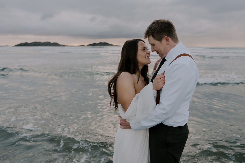 Couple embracing and smiling on a beach with gentle waves and cloudy sky. She wears a white dress, he a white shirt with suspenders. Romantic mood.  Vancouver Moody Romantic intimate Wedding Photographer.