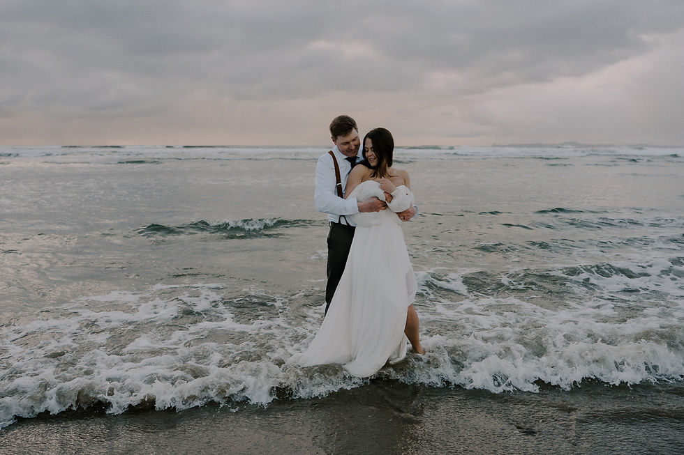 A couple in formal attire embraces at the shoreline, waves gently lapping. Cloudy sky sets a serene, romantic mood.  Vancouver Moody Romantic intimate Wedding Photographer.