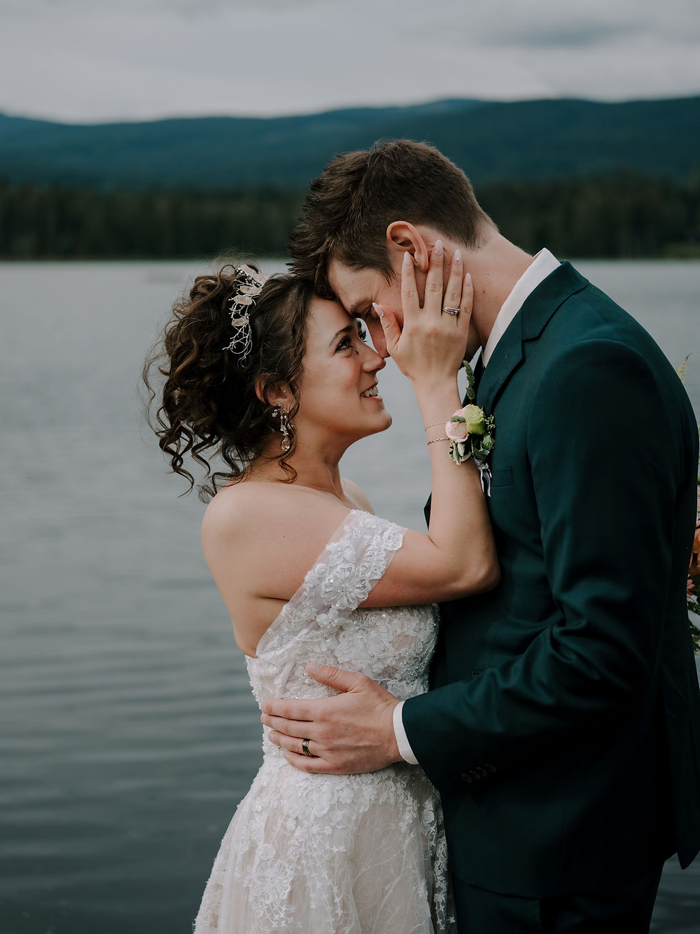 Bride and groom embrace by a lake, foreheads touching. She's in a lace dress, he's in a dark suit. Romantic, serene backdrop with mountains. Dark and moody romantic wedding Vancouver photographer.