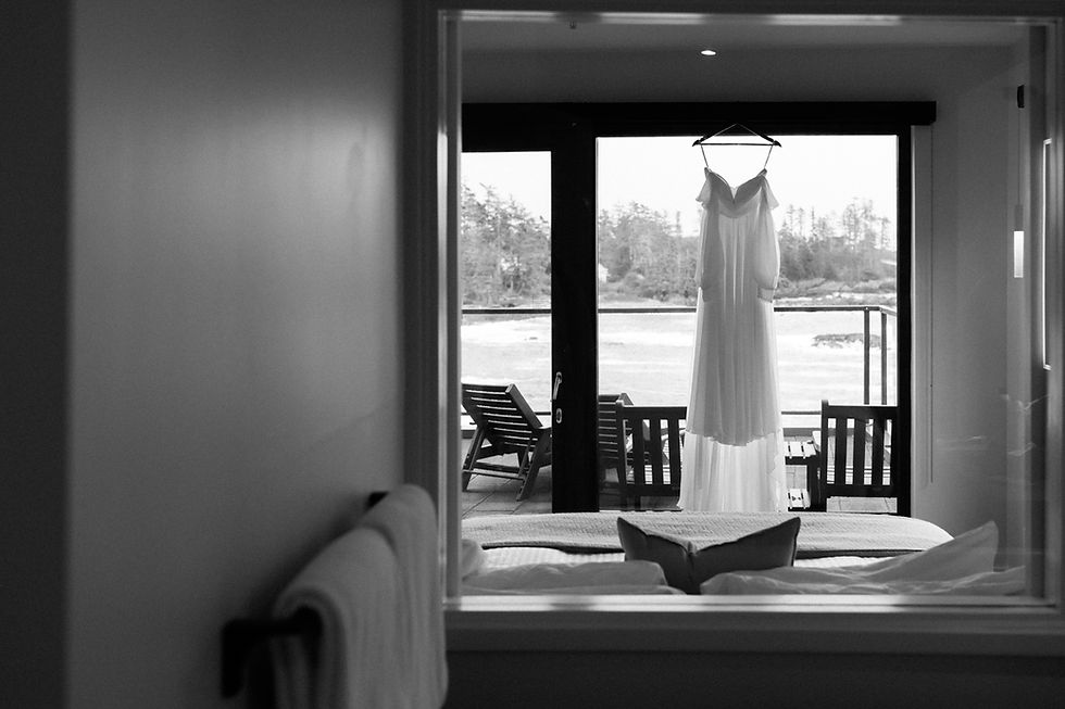 White dress hangs in a bright room with a view of chairs on a patio and trees outside. Towels are on a rack, creating a serene mood.  Vancouver Moody Romantic intimate Wedding Photographer.