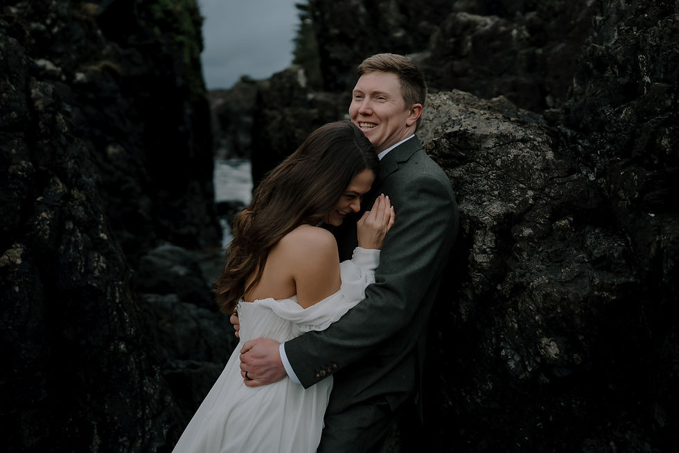 Couple embracing on rocky coastline; woman in white dress, man in dark suit. Moody, overcast sky in background, expressing happiness.  Vancouver Moody Romantic intimate Wedding Photographer.