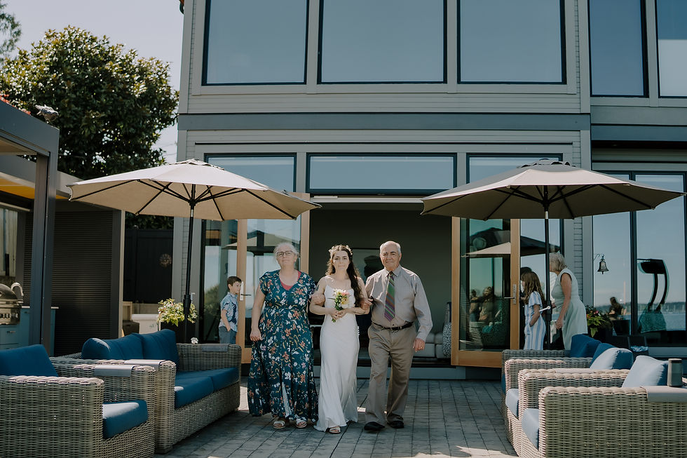Bride escorted by older couple across a sunny patio at a modern house, with wicker seating and beige umbrellas. Moody and intimate Vancouver wedding photographer with a dark and heartfelt signature look. Small backyard wedding documented.