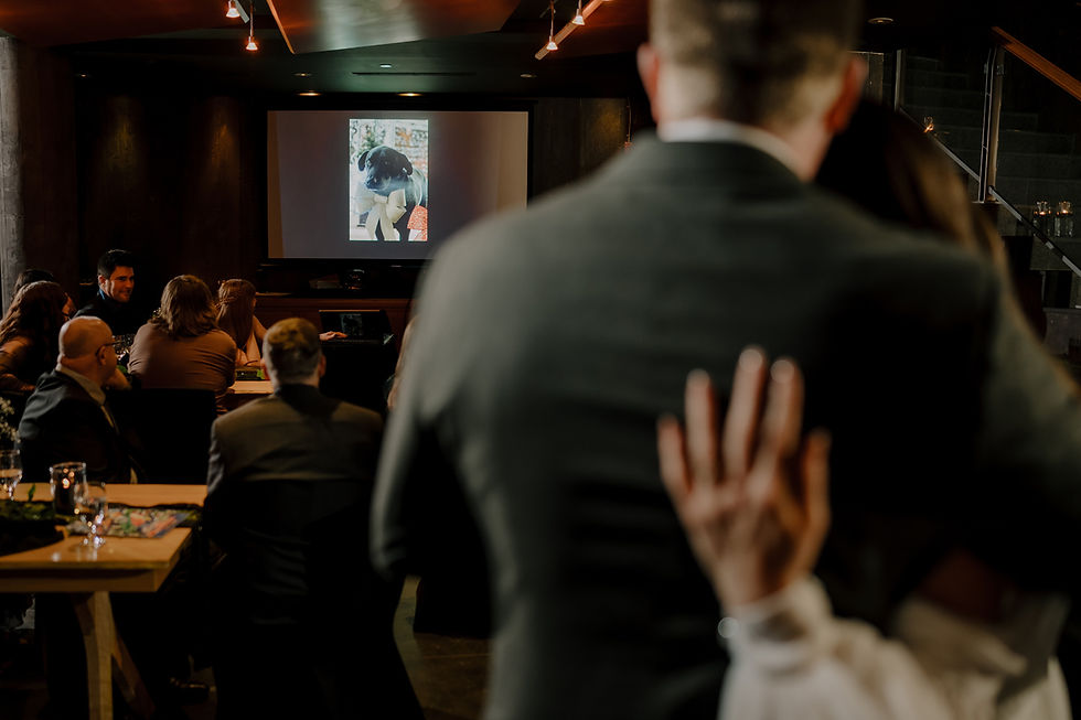 Couple embracing at an event, watching a slideshow featuring a dog on a screen. Dimly lit room with guests seated at tables.  Vancouver Moody Romantic intimate Wedding Photographer.