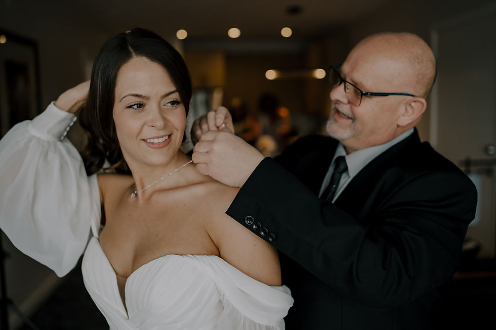 A man helps a woman in a white dress with her necklace. They smile in a warmly lit room, creating a joyful atmosphere.  Vancouver Moody Romantic intimate Wedding Photographer.