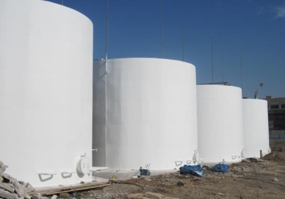 Four large white cylindrical tanks stand in a sandy area under a clear blue sky, with some debris and tarps nearby. showcasing best thermal insulation for tanks