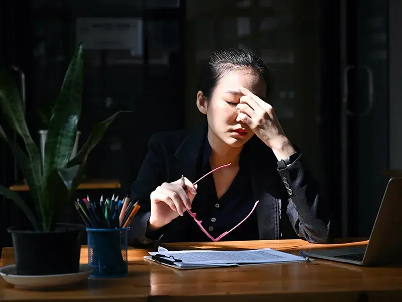 A woman in a dark suit, sitting at a desk with a laptop, holding glasses and rubbing their eyes. Sunlit room, plant and colorful pencils nearby. Suffering from Chronic Fatigue