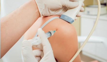 A medical professional uses an ultrasound probe and needle for a procedure on a patient's knee, wearing gloves in a clinical setting.