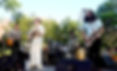 Outdoor band performs on a park stage, with guitars, drums, and microphones under trees on a sunny day.
