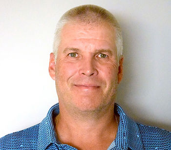 Smiling middle-aged man with short gray hair in a blue patterned shirt, posed against a plain white background.