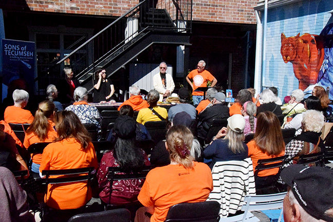 Outdoor talk with seated audience facing speakers at a brick courtyard, with Sons of Tecumseh sign and horse mural.