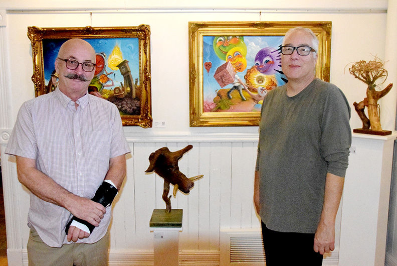 Two men stand in an art gallery beside colorful framed paintings and a sculpture.
