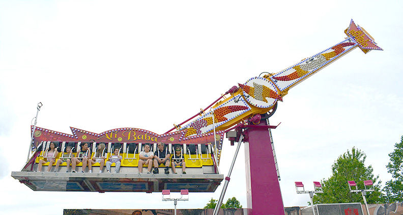 Amusement park ride labeled Ali Baba swings with riders seated in a bright yellow gondola against a cloudy sky.