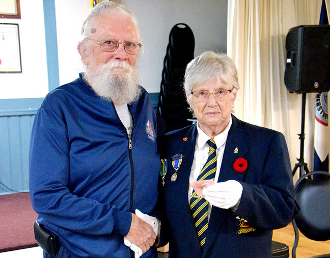 An elderly man and woman shake hands indoors; she wears medals, a poppy, and a striped tie, with a speaker and flag behind.