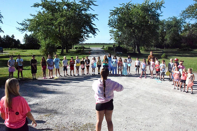 Children and a few adults stand in a circle on a gravel path outdoors, with two women leading in a sunny park.