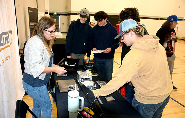 People browse a trade-show booth in a gym, while a woman arranges items on a table and others inspect papers and products.