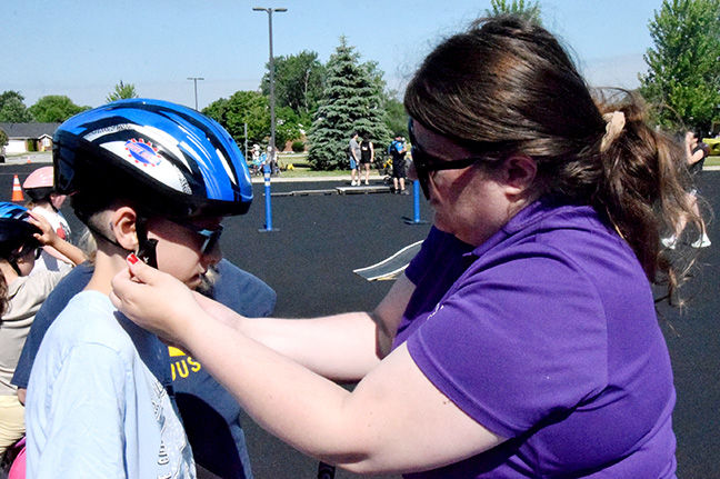 Woman in purple adjusts a boy’s blue helmet strap in a sunny parking lot, with other people and trees in the background.