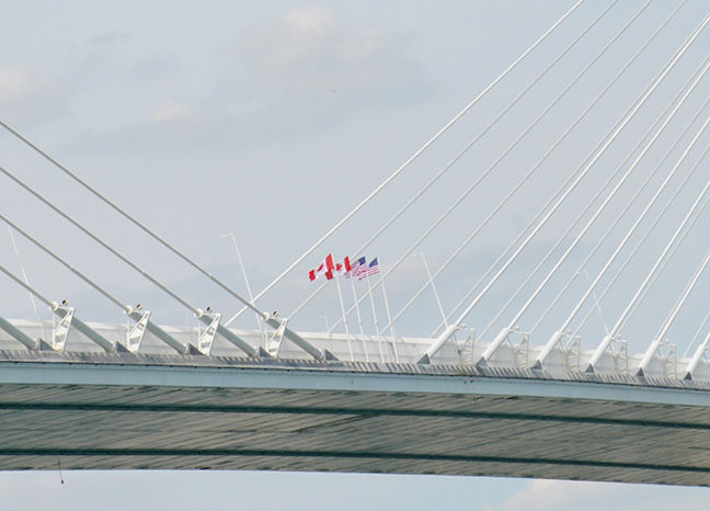 Suspension bridge deck with white cables and Canada and U.S. flags against a cloudy sky.