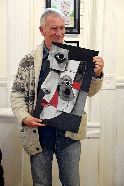 Man holding abstract black, white, and red artwork, smiling in a cozy room with white walls and framed pictures. Wearing patterned sweater.