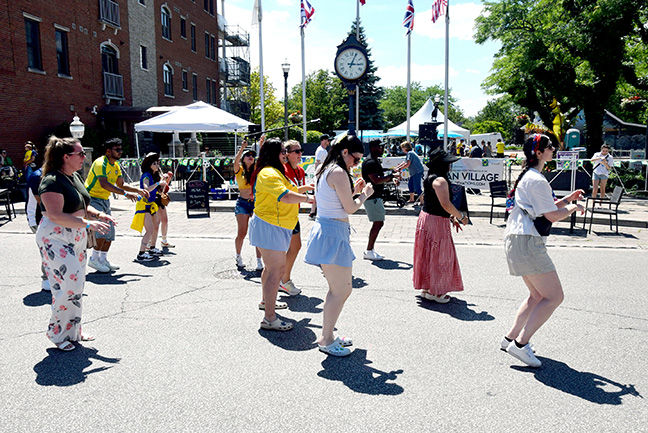 People dance in a sunny street festival by a clock tower and tents, with a sign reading AN VILLAGE in the background.