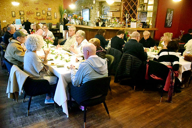 People dining at a restaurant, sitting at tables with white tablecloths, in a cozy setting with a bar in the background. Warm lighting.