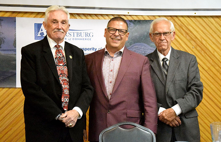 Three men in suits stand smiling at a business event, with an Amherstburg Chamber of Commerce banner behind them.