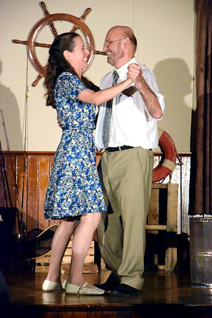 Smiling couple dances on a small stage, holding hands, with a ship’s wheel and life preserver behind them.