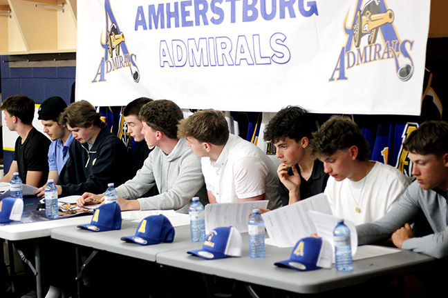 Young men sit at tables signing papers beneath an Amherstburg Admirals banner, with blue caps and water bottles in front.