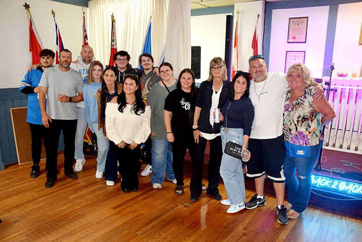 Group of smiling adults posing indoors on a wooden floor, with flags, framed certificates, and a neon BACK 2 BACK sign in back.