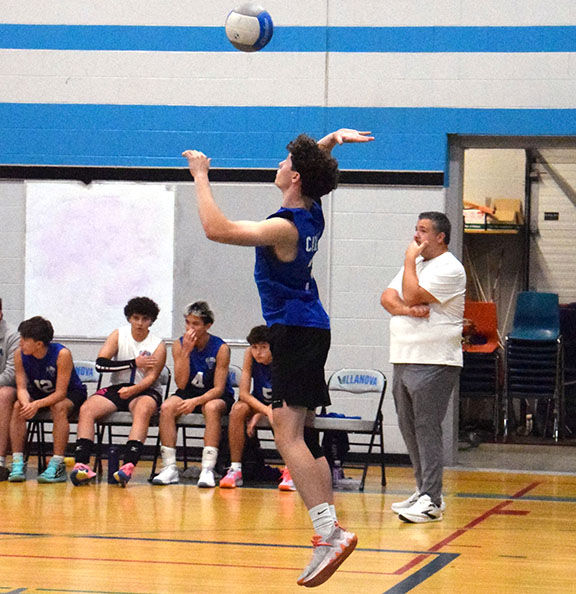 Indoor volleyball practice: a player in blue jumps to hit a ball while teammates and a coach watch in the gym.