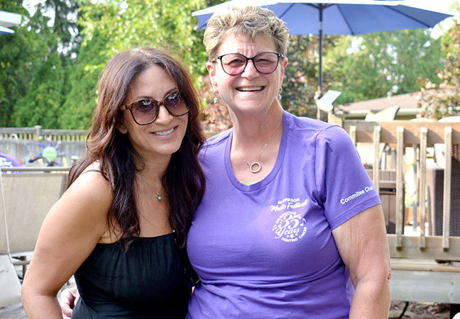Two smiling women pose outdoors on a deck, one in a purple shirt with 35 Years text, trees and a patio umbrella behind.