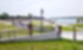Waterfront park with curving paths, black railings, lamp posts, green lawns, and calm lake under a bright sky.