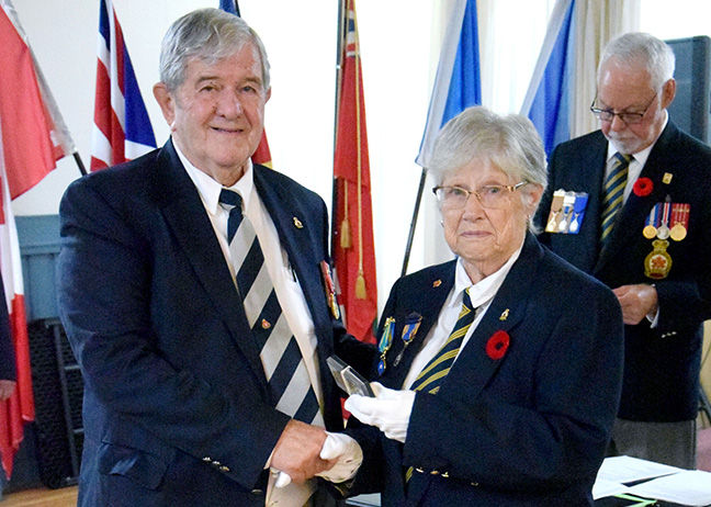Two elderly veterans in navy blazers shake hands, one holding a plaque, with flags and another medal-wearing man behind.