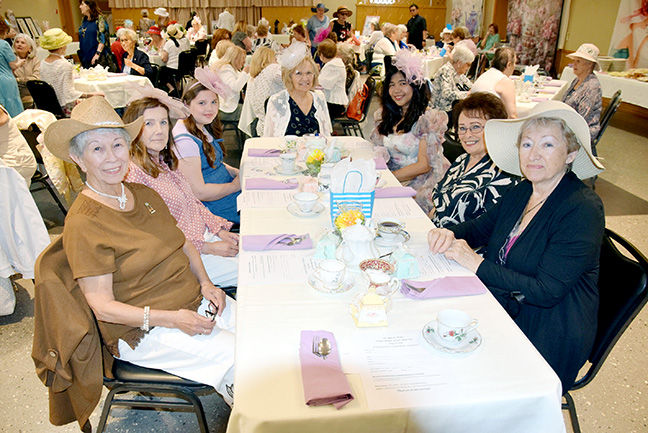 Group of women in hats sit smiling at a banquet table in a bright hall, with pastel napkins and cups at a lively indoor event.