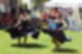 Two dancers in black dresses twirl barefoot on grass at an outdoor festival, with tents, art displays, and spectators behind.
