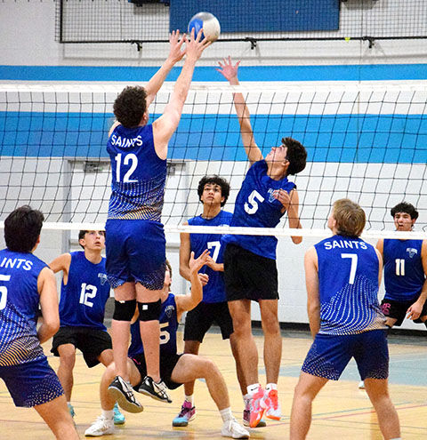 Volleyball players in blue SAINTS jerseys jump at the net for a block and spike in an indoor gym.