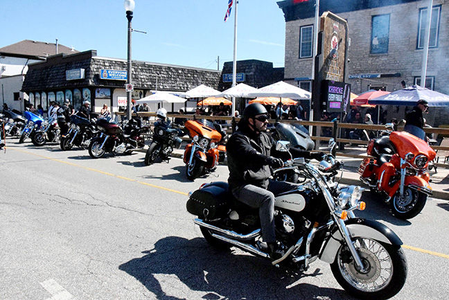 Motorcyclists line a sunny street outside a waterfront bar, with a rider on a black cruiser in the foreground.