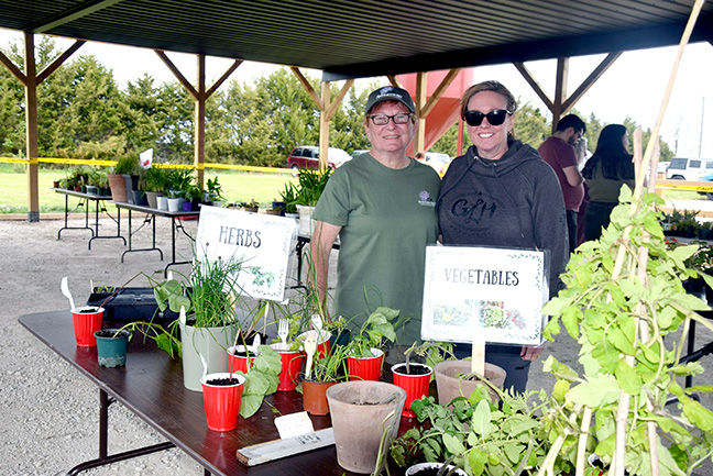 Two people smiling at a plant sale under a pavilion. Tables display herbs and vegetables in pots. Signs read "HERBS" and "VEGETABLES."