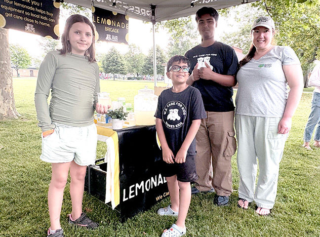 Four smiling people stand at a park lemonade stand with Lemonade Brigade signs and a pitcher on the table.