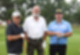 Three men pose on a golf course holding clubs, wearing white, black, and blue shirts, with trees in the background.